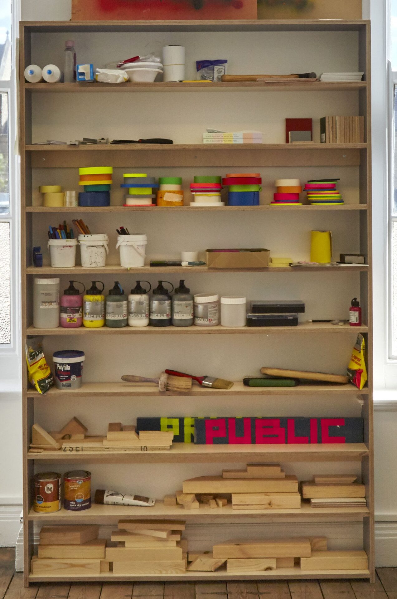 A tall studio shelving unit filled with paint pots, coloured tape, brushes and cut timber — a working set of materials.